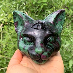 May include: A carved stone cat head figurine featuring a third eye on the forehead. The sculpture is crafted from a mottled mineral with deep green, black, and dark magenta-red patches, polished to a smooth, glossy finish with pointed ears.