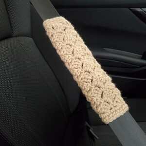 May include: A beige, crocheted seat belt cover is shown in the image. The cover is made of a soft, textured yarn and is designed to fit over a standard seat belt. The cover is shown in a car interior, resting on a gray seat belt.