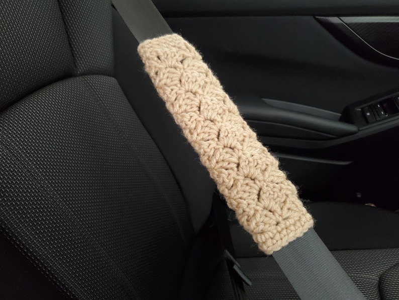 May include: A beige, crocheted seat belt cover is shown in the image. The cover is made of a soft, textured yarn and is designed to fit over a standard seat belt. The cover is shown in a car interior, resting on a gray seat belt.