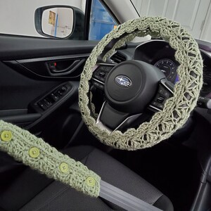 Op de afbeelding: Een saliegroene gehaakte stuurhoes en een bijpassende veiligheidsgordelhoes met vier kleine ronde knopen. De accessoires zijn gemaakt met een opengewerkt garenpatroon en zijn bevestigd aan het stuur en de veiligheidsgordel van een voertuig.
