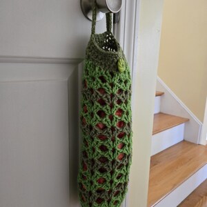 Puede incluir: Una bolsa de crochet verde con un bot&oacute;n colgando de un pomo de puerta. La bolsa est&aacute; hecha con un patr&oacute;n repetitivo de hilo verde y marr&oacute;n.