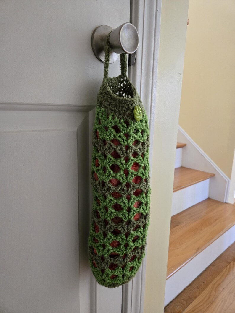 Puede incluir: Una bolsa de crochet verde con un bot&oacute;n colgando de un pomo de puerta. La bolsa est&aacute; hecha con un patr&oacute;n repetitivo de hilo verde y marr&oacute;n.