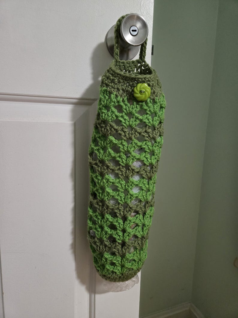 Puede incluir: Una bolsa de crochet verde y marr&oacute;n para guardar bolsas de pl&aacute;stico de la compra. La bolsa est&aacute; colgada de una puerta blanca con un pomo de puerta plateado. Un bot&oacute;n verde est&aacute; sujeto a la bolsa.