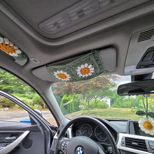 May include: A set of three crochet car accessories in sage green, white, and yellow. The set includes a handle cover, a sun visor cover featuring two daisy motifs, and a hanging rearview mirror ornament shaped like a single daisy flower.