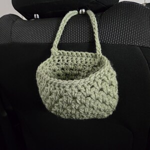Op de afbeelding: Een saliegroene gehaakte hangmand met &eacute;&eacute;n handvat, gemaakt van dik garen in een gestructureerd steekpatroon. De mand is ontworpen om aan een steun te worden opgehangen en heeft een open bovenkant en een rond, diep opbergcompartiment.
