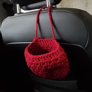Op de afbeelding: Een rode gehaakte hangende mand met een geweven textuur, hangend aan de hoofdsteun van een autostoel.