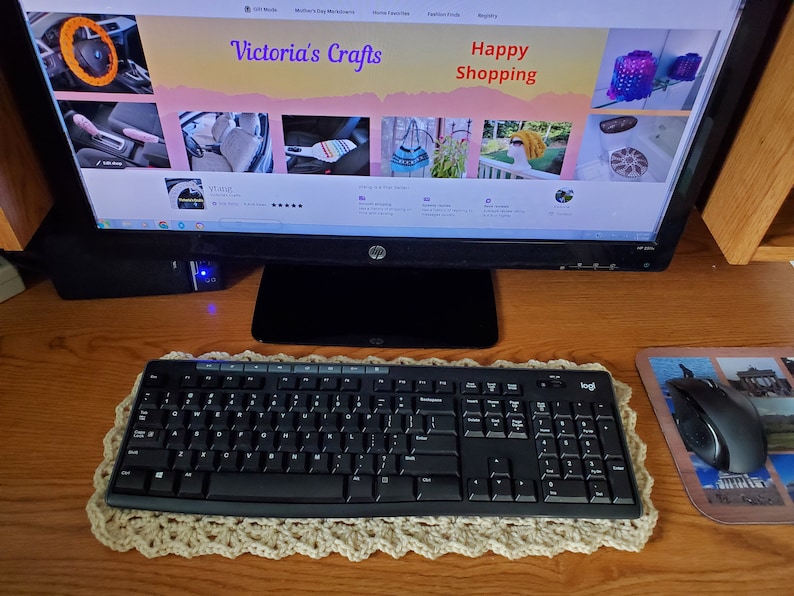 May include: A computer monitor displaying a website called "Victoria's Crafts" with the text "Happy Shopping" on the top. The website features images of various handmade crafts, including a steering wheel cover, car seat covers, and crocheted items. A black keyboard and mouse are on a wooden desk in front of the monitor.