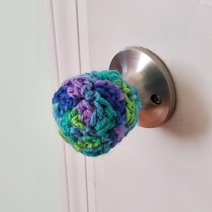 May include: A crocheted door knob cover in shades of blue, green, and purple. The cover is made with a textured stitch pattern.