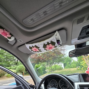 May include: A crochet car interior accessory set featuring a white, dark green, and pink floral granny square pattern. The set includes a cover for the passenger grab handle and a matching visor organizer sleeve, plus a hanging pink flower charm.