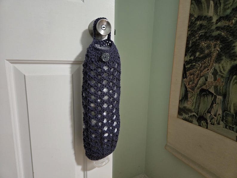 Puede incluir: Una bolsa de crochet azul oscuro con cierre de cord&oacute;n cuelga de un pomo de puerta. La bolsa est&aacute; hecha de un delicado patr&oacute;n de tejido abierto y est&aacute; dise&ntilde;ada para contener art&iacute;culos de comestibles.