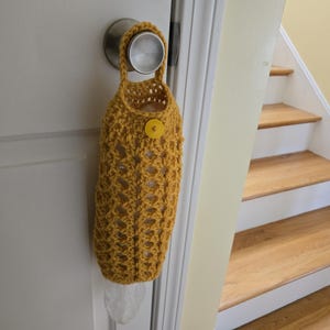 May include: A crocheted yellow plastic bag holder with a yellow button hanging on a white door.