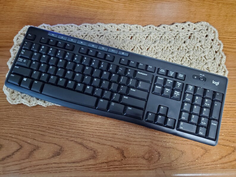May include: A black Logitech wireless keyboard with a number pad and a white logo. The keyboard is on a beige crocheted surface.