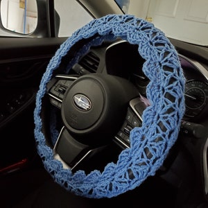May include: A blue crocheted steering wheel cover with a delicate floral pattern. The cover is made of soft yarn and is designed to fit most standard steering wheels.