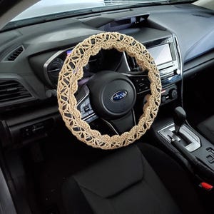 May include: A beige, crocheted steering wheel cover is installed on a black steering wheel in a car. The cover has a geometric pattern and adds a decorative touch to the vehicle's interior. The car's dashboard and seats are also visible.