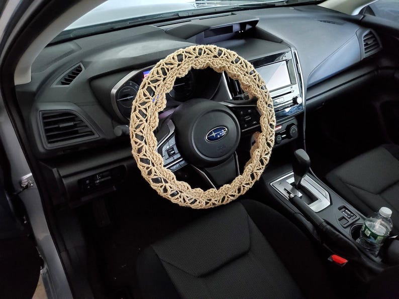 May include: A beige, crocheted steering wheel cover is installed on a black steering wheel in a car. The cover has a geometric pattern and adds a decorative touch to the vehicle's interior. The car's dashboard and seats are also visible.