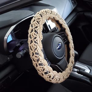 May include: A beige crochet steering wheel cover is shown in a car interior. The cover is made of a light-colored yarn with an open, lacy design. The steering wheel is black, and the car's dashboard and interior are also black.