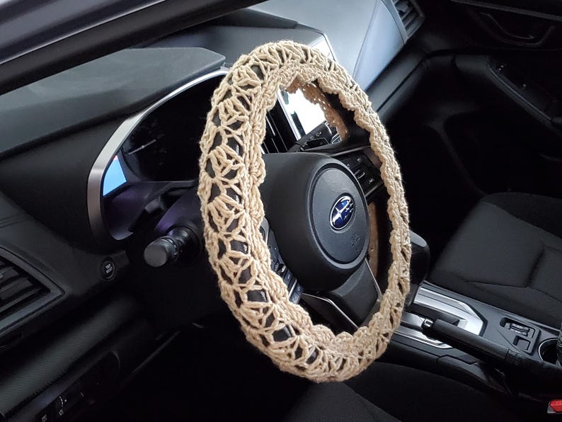 May include: A beige crochet steering wheel cover is shown in a car interior. The cover is made of a light-colored yarn with an open, lacy design. The steering wheel is black, and the car's dashboard and interior are also black.