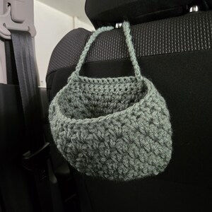 Op de afbeelding: Een saliegroene gehaakte hangmand met een gestructureerd steekpatroon en een enkele handgreep, ontworpen om aan een hoofdsteun te hangen. De mand heeft een afgeronde, open komvorm, vervaardigd uit dik garen in een gedempte groene tint.
