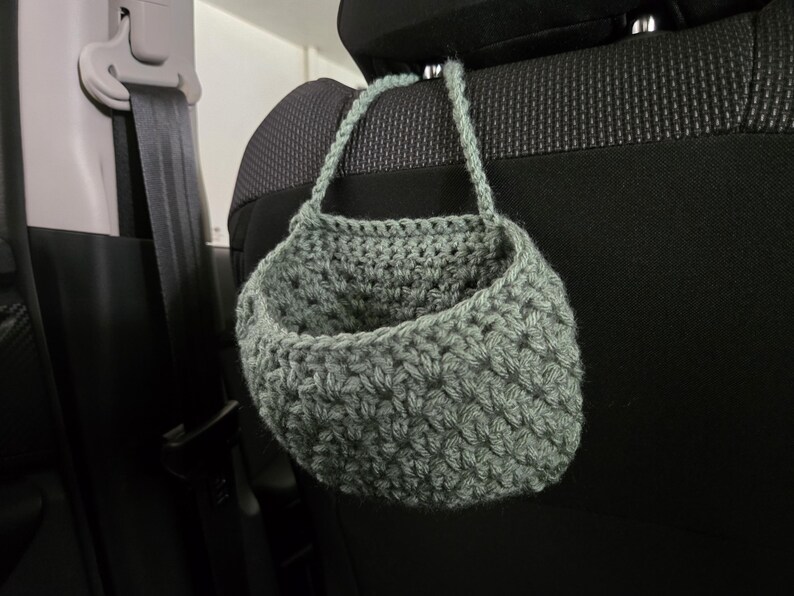 Op de afbeelding: Een saliegroene gehaakte hangmand met een gestructureerd steekpatroon en een enkele handgreep, ontworpen om aan een hoofdsteun te hangen. De mand heeft een afgeronde, open komvorm, vervaardigd uit dik garen in een gedempte groene tint.