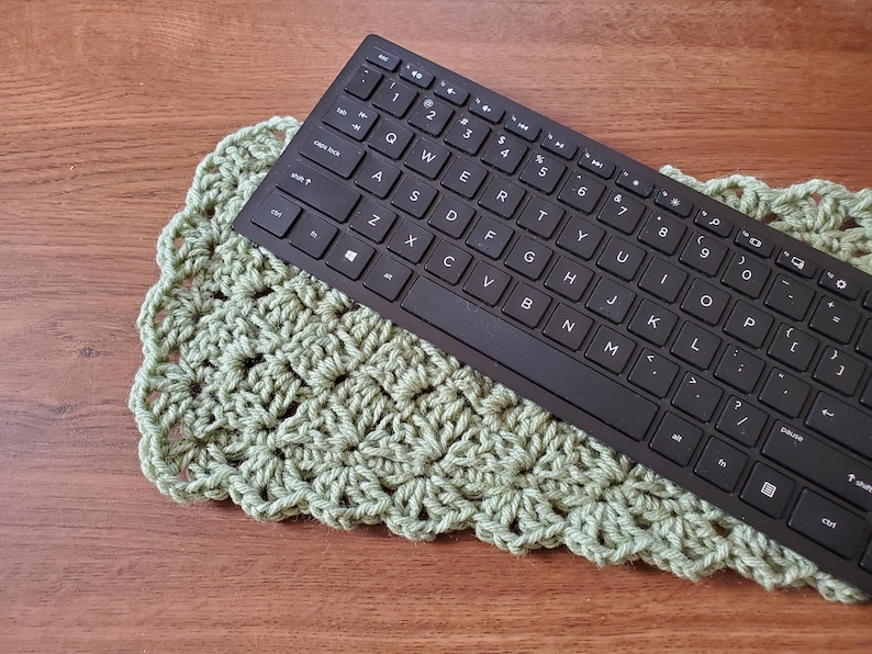 May include: A black wireless keyboard with a green crocheted keyboard mat. The keyboard has a standard layout with keys for numbers, letters, and punctuation. The mat is made of a soft, textured yarn.