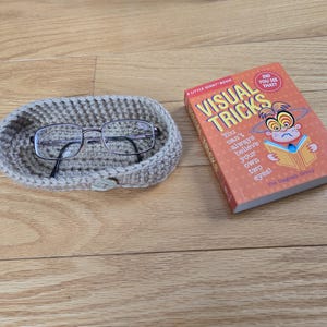 May include: A beige crocheted eyeglass holder with a pair of glasses inside. The holder is sitting on a wooden surface next to a book titled "Visual Tricks" with a cartoon monkey on the cover. The book says "You can't always believe your own two eyes!"