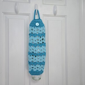 May include: A crocheted blue and light blue plastic bag holder with a button closure hanging on a white door.
