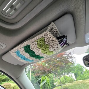 May include: A beige crochet car visor organizer featuring a granny square pattern with accents of light blue, dark green, and lime green yarn. The rectangular sleeve is attached to a vehicle sun visor and holds a pair of dark-framed sunglasses.