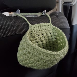 Op de afbeelding: Een saliegroene gehaakte hangmand met een brede, open bovenkant en een stevige garenlus om op te hangen. De mand is gemaakt met een gestructureerd steekpatroon en is ontworpen om aan een steunpaal of haak te hangen via het ge&iuml;ntegreerde handvat.