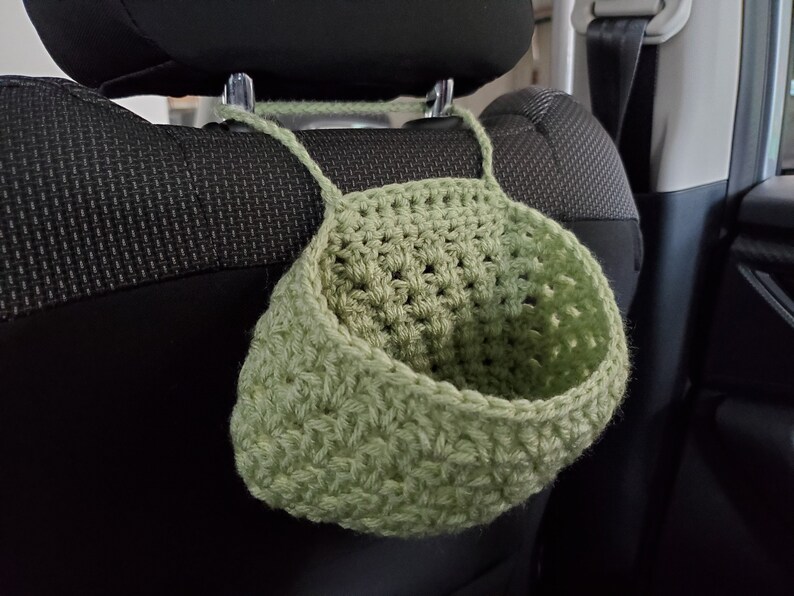 Op de afbeelding: Een saliegroene gehaakte hangmand met een brede, open bovenkant en een stevige garenlus om op te hangen. De mand is gemaakt met een gestructureerd steekpatroon en is ontworpen om aan een steunpaal of haak te hangen via het ge&iuml;ntegreerde handvat.
