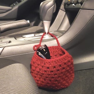 Op de afbeelding: Een rode gehaakte hangende organizer met een zonnebril erin, hangend aan de versnellingspook in een auto.