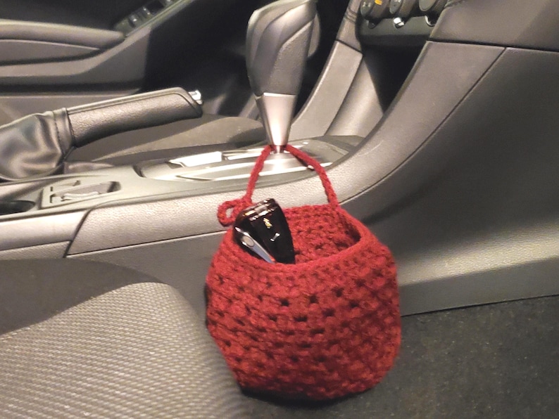 Op de afbeelding: Een rode gehaakte hangende organizer met een zonnebril erin, hangend aan de versnellingspook in een auto.