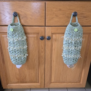 May include: Two green crocheted bags with white trim hang from hooks on a wooden cabinet. Each bag has a green button.
