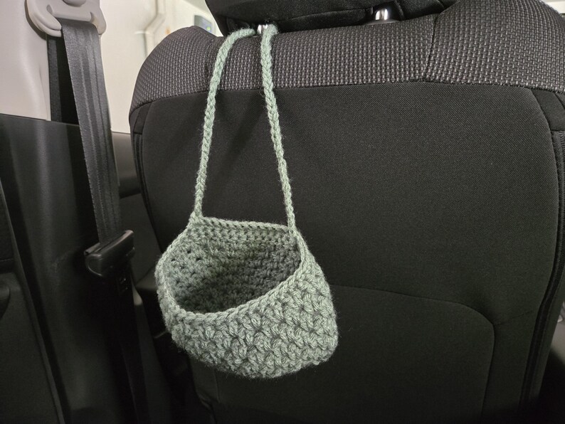 Op de afbeelding: Een saliegroene gehaakte hangmand met een gestructureerd steekpatroon en een enkele ge&iuml;ntegreerde lus. De mand is gemaakt van dik garen en heeft een brede, open bovenkant, ontworpen om aan een haak of steunstructuur te worden opgehangen.