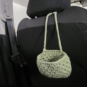 Op de afbeelding: Een saliegroene gehaakte hangmand met &eacute;&eacute;n hengsel, opgehangen aan een metalen haak bevestigd aan de hoofdsteun van een zwarte autostoel. De mand heeft een gestructureerd, open steekpatroon en een afgeronde, komvormige basis voor opslag.