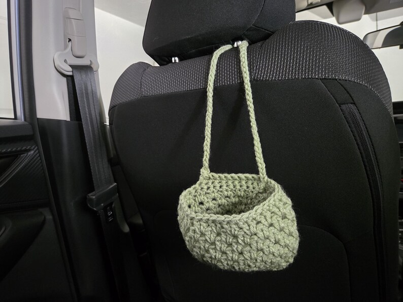Op de afbeelding: Een saliegroene gehaakte hangmand met &eacute;&eacute;n hengsel, opgehangen aan een metalen haak bevestigd aan de hoofdsteun van een zwarte autostoel. De mand heeft een gestructureerd, open steekpatroon en een afgeronde, komvormige basis voor opslag.