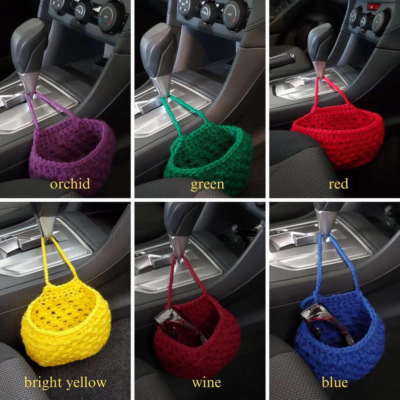 Op de afbeelding: Zes gehaakte hangmanden in verschillende kleuren, waaronder orchidee, groen, rood, felgeel, wijnrood en blauw. De manden hangen aan de versnellingspook van een auto.