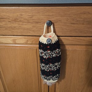 Puede incluir: Un porta bolsas de crochet con rayas blancas y negras y una parte superior de crochet beige. El porta bolsas est&aacute; colgado en una puerta de armario de madera con un pomo negro.