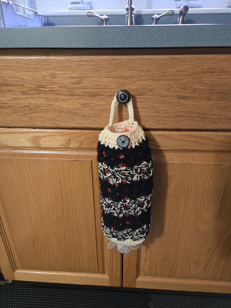 Puede incluir: Un porta bolsas de crochet con rayas blancas y negras y una parte superior de crochet beige. El porta bolsas est&aacute; colgado en una puerta de armario de madera con un pomo negro.