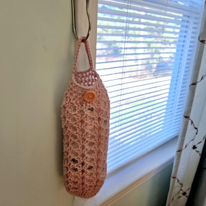 May include: A pink crocheted bag with a button closure hangs on a hook near a window. The bag is made of a delicate, open-weave pattern.