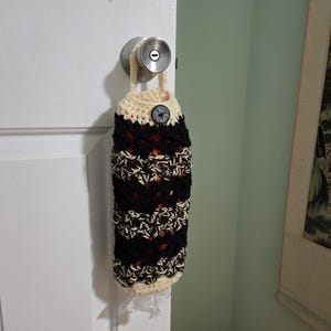 Puede incluir: Una bolsa de ganchillo con rayas negras, blancas y rojas colgada de un pomo de puerta. La bolsa tiene un cierre de bot&oacute;n y un ribete de ganchillo blanco.