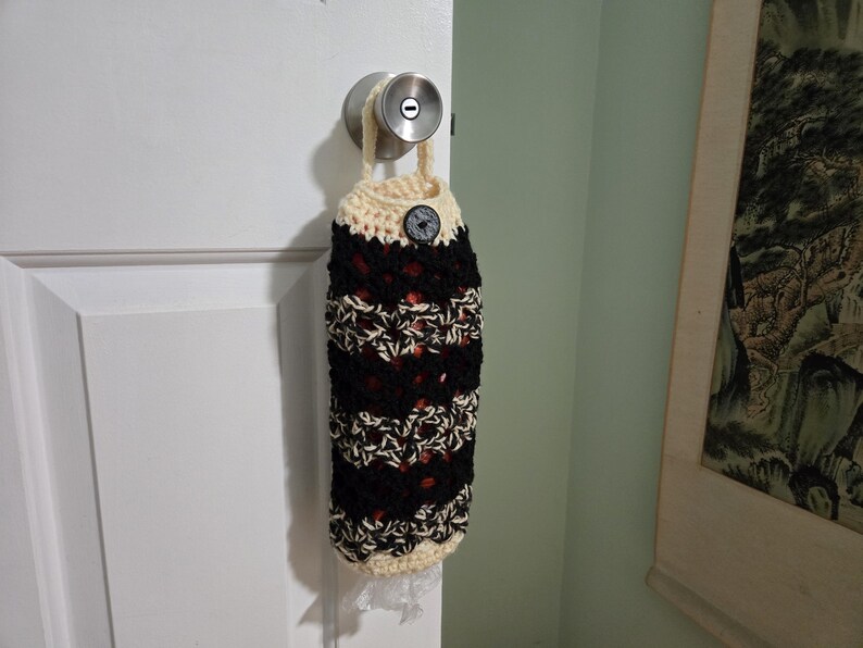 Puede incluir: Una bolsa de ganchillo con rayas negras, blancas y rojas colgada de un pomo de puerta. La bolsa tiene un cierre de bot&oacute;n y un ribete de ganchillo blanco.