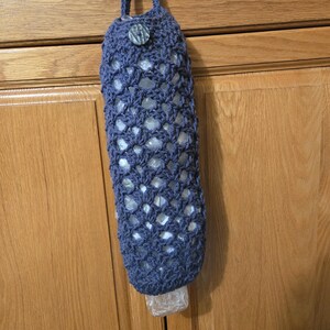 Puede incluir: Una bolsa de crochet azul para guardar bolsas de pl&aacute;stico de la compra cuelga del pomo de una puerta de armario. La bolsa tiene un bot&oacute;n de madera y un dise&ntilde;o de crochet.