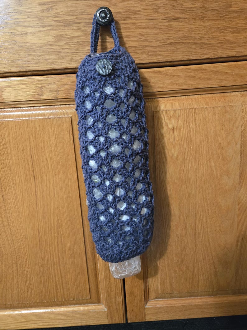 Puede incluir: Una bolsa de crochet azul para guardar bolsas de pl&aacute;stico de la compra cuelga del pomo de una puerta de armario. La bolsa tiene un bot&oacute;n de madera y un dise&ntilde;o de crochet.
