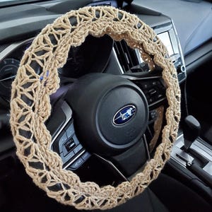 May include: A beige crochet steering wheel cover, shown installed in a car. The cover has an open, lacy design and is wrapped around the black steering wheel. The car's dashboard and other interior details are visible in the background.