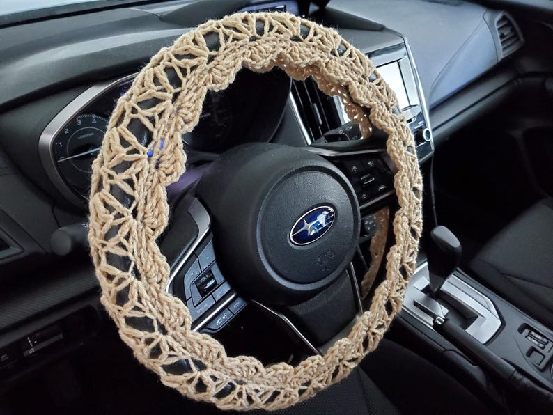 May include: A beige crochet steering wheel cover, shown installed in a car. The cover has an open, lacy design and is wrapped around the black steering wheel. The car's dashboard and other interior details are visible in the background.
