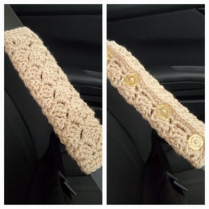 May include: A beige, crocheted seat belt cover. The cover is rectangular and has a textured pattern. Three clear buttons secure the cover. The cover is designed to fit over a car seat belt, providing comfort and style.