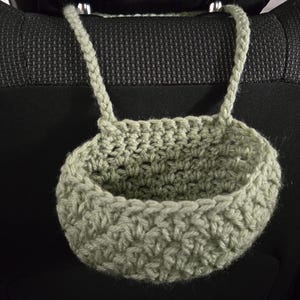 Op de afbeelding: Een saliegroene gehaakte hangmand met een ronde bodem en een gestructureerd steekpatroon. De mand is voorzien van twee gevlochten handvatten aan de bovenrand, waardoor deze aan een steun kan worden opgehangen voor opslag of organisatie.