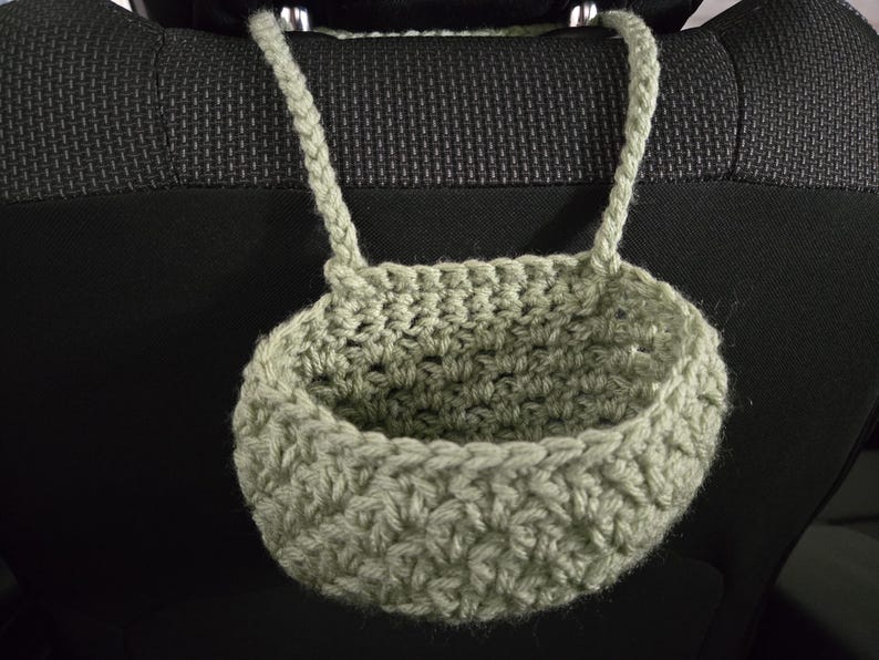 Op de afbeelding: Een saliegroene gehaakte hangmand met een ronde bodem en een gestructureerd steekpatroon. De mand is voorzien van twee gevlochten handvatten aan de bovenrand, waardoor deze aan een steun kan worden opgehangen voor opslag of organisatie.