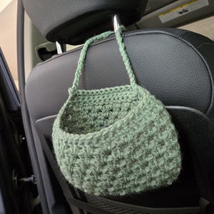 Op de afbeelding: Een saliegroene gehaakte hangmand met een gestructureerd steekpatroon en een enkele handgreep, opgehangen aan de metalen hoofdsteun van een zwarte autostoel. De mand heeft een open bovenkant en is gemaakt van dik garen.