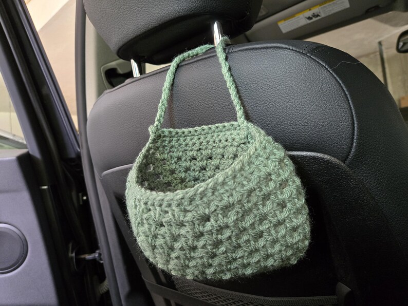 Op de afbeelding: Een saliegroene gehaakte hangmand met een gestructureerd steekpatroon en een enkele handgreep, opgehangen aan de metalen hoofdsteun van een zwarte autostoel. De mand heeft een open bovenkant en is gemaakt van dik garen.